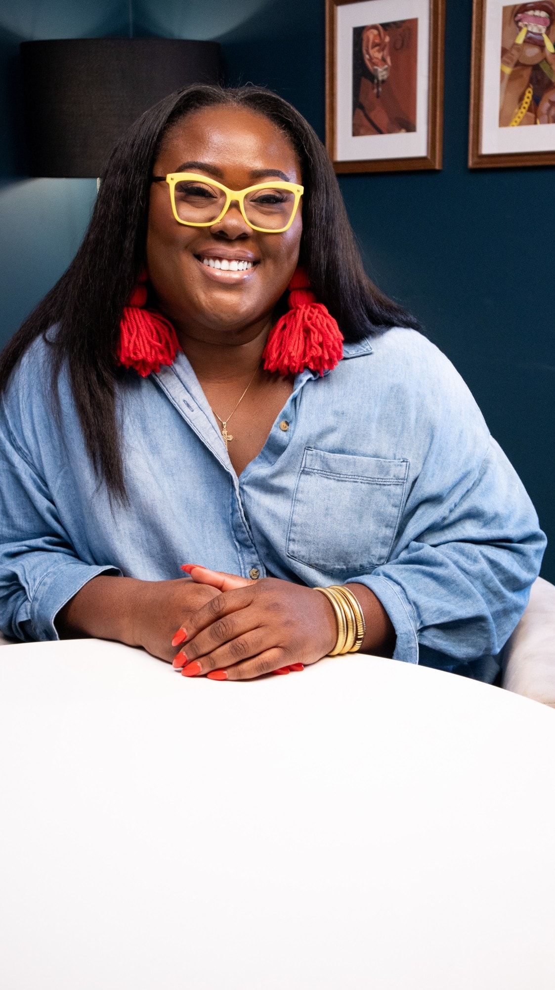 Ona smiling at a table in a denim shirt, yellow glasses and red tassel earrings