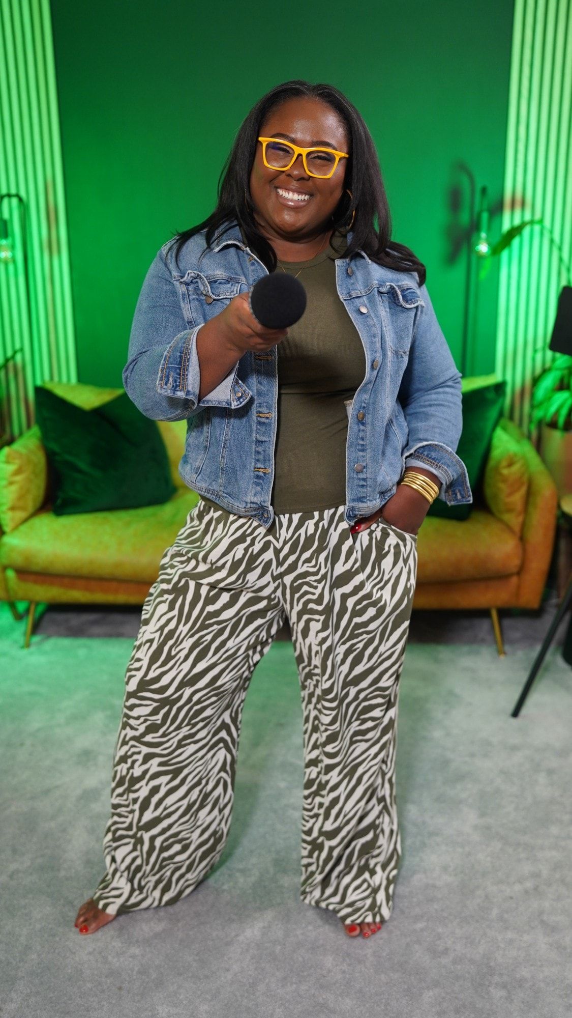 Ona smiling in yellow glasses and a denim jacket, holding a microphone out toward the camera